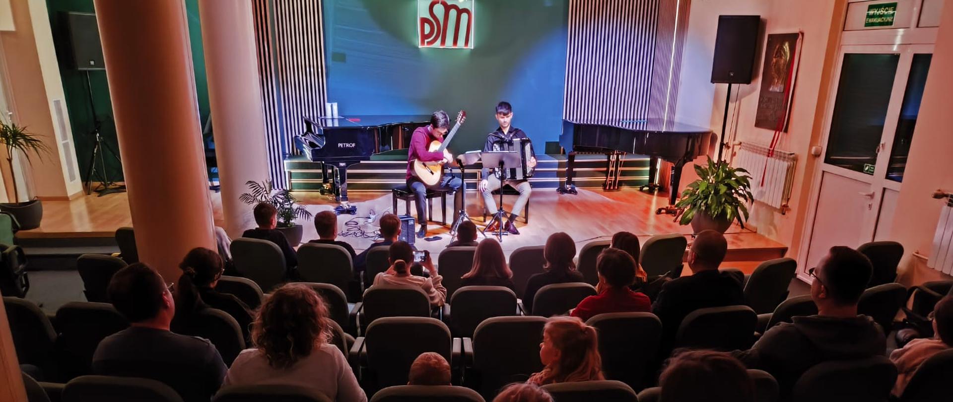 Na zdjęciu widoczna jest oświetlona reflektorami scena sali koncertowej w szkole muzycznej na której grają artyści na gitarze jako akordeonie siedzący na krzesłach za nimi znajduje się ściana w zielonym kolorze z logo szkoły z elementami akustycznymi. Na dole zdjęcia widoczna jest publiczność która podziwia występ artystów