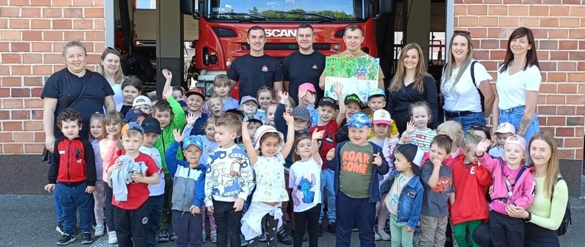 Na zdjęciu znajdują się dzieci wraz z opiekunami oraz strażacy . Zdjęcie jest zrobione na tle samochodu pożarniczego