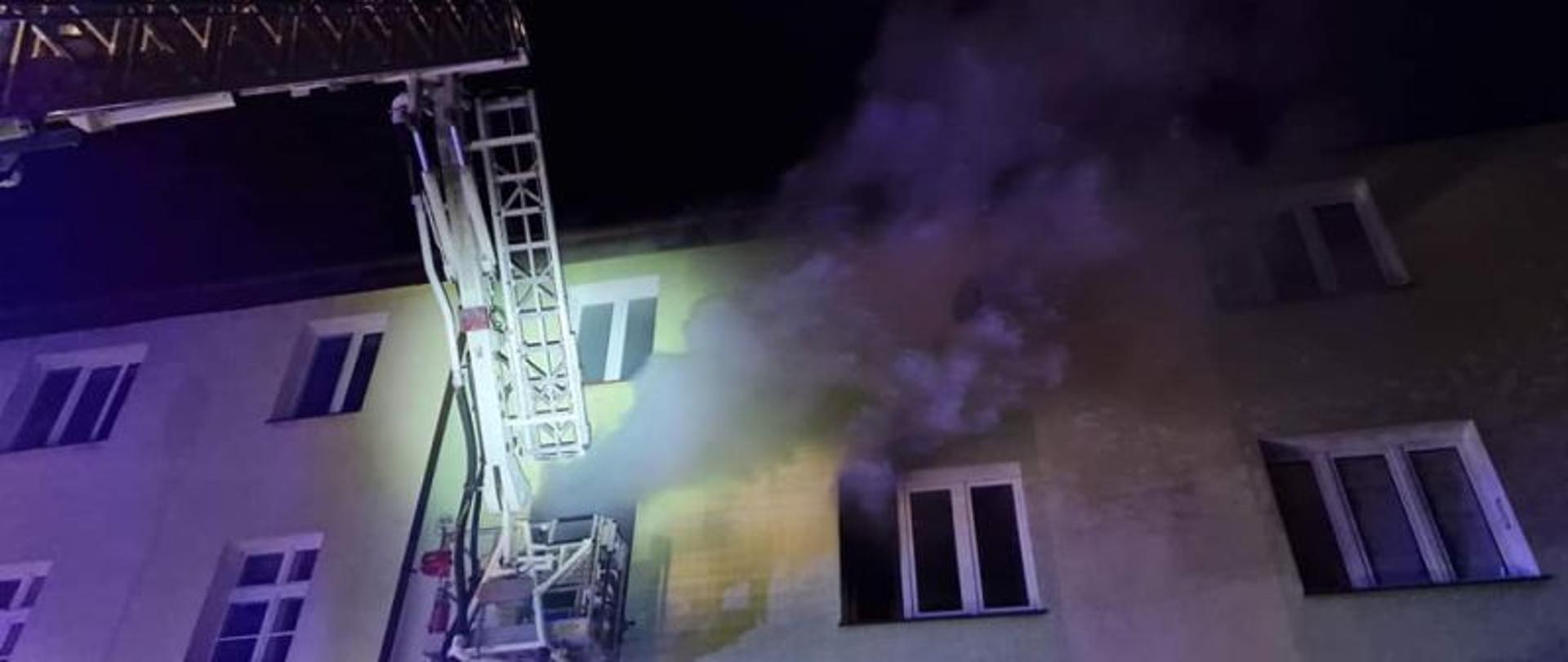 Zdjęcie przedstawia rozstawione ramię podnośnika skierowane w stronę ściany budynku i okna z którego wydobywa się dym. Tło stanowi czarne, nocne niebo. Ściana i ramię oświetlona sztucznym światłem latarek i najaśnic.