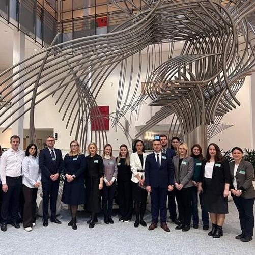 18 osób pozuje do zdjęcia w hallu budynku z dużą aluminiową rzeźbą w tle.