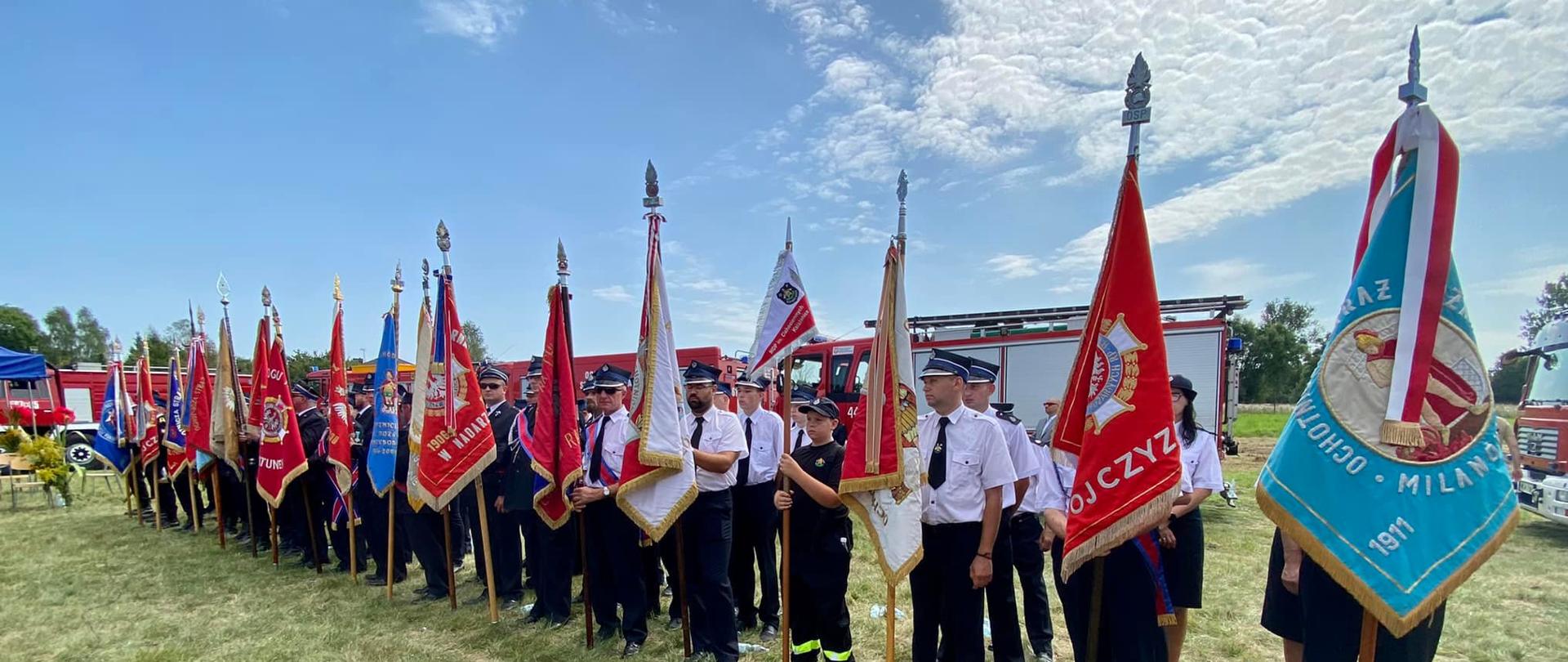 Poczty sztandarowe ustawione w szeregu w trakcie uroczystej mszy świętej. W tle widać samochody pożarnicze.
