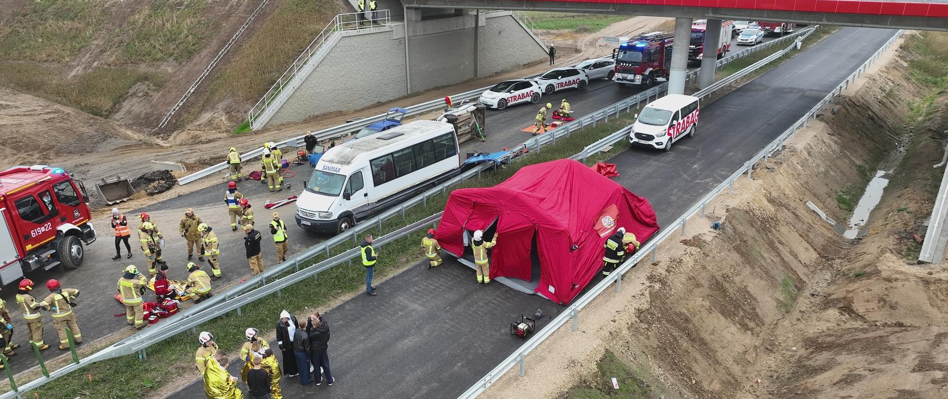 Na nowej drodze ekspresowej trwa akcja ratunkowa po wypadku. Widać strażaków, karetki, wozy strażackie i biały bus. Na jezdni stoi czerwony namiot ratowniczy, a ratownicy pomagają poszkodowanym.