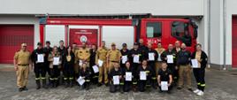 Zdjęcie przedstawia grupowo uczestników szkolenia trzymających w ręce dyplomy na tle samochodu pożarniczego.
