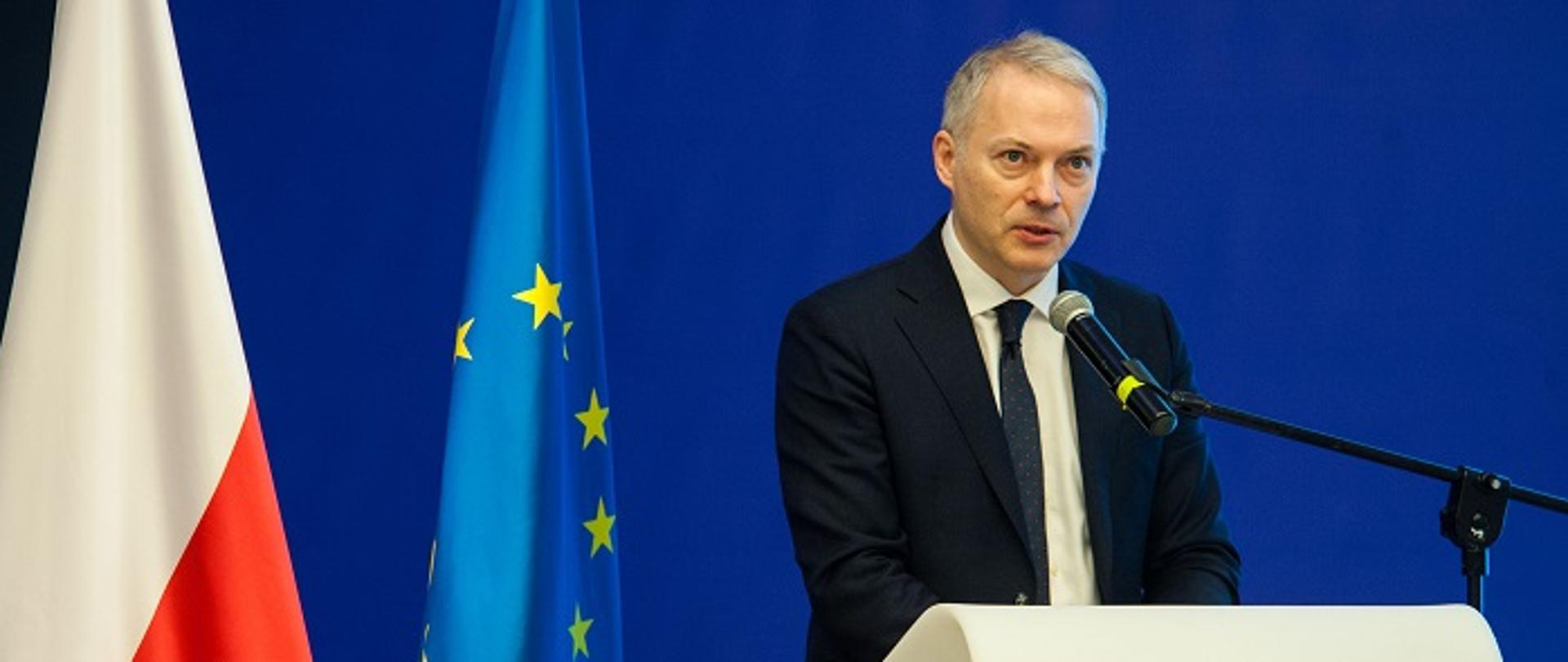 Wiceminister Jacek Żalek stoi przy mównicy, przed mikrofonem, za nim flagi
Polski i UE.