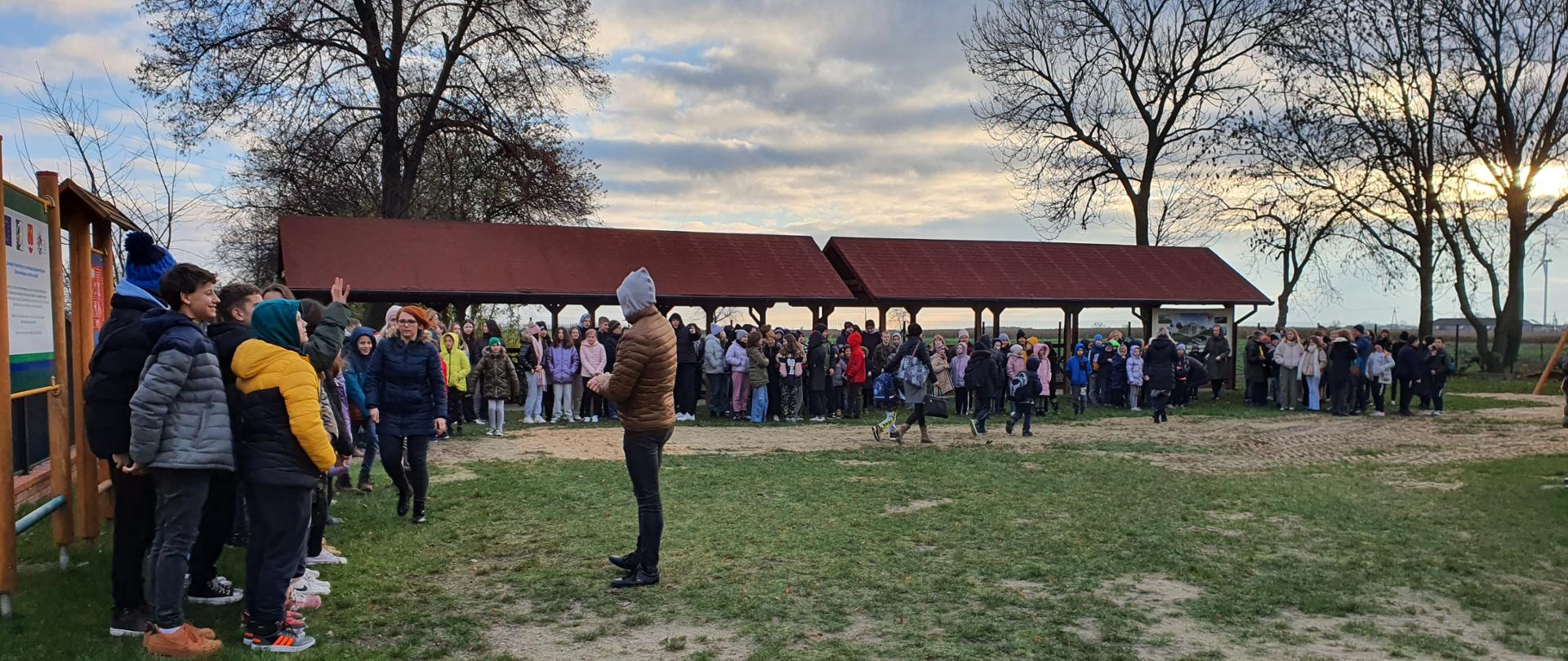 Zdjęcie przedstawia dzieci i młodzież szkolną zgromadzoną na terenie zielony, pod wiatami. W tle widoczne tereny zielone