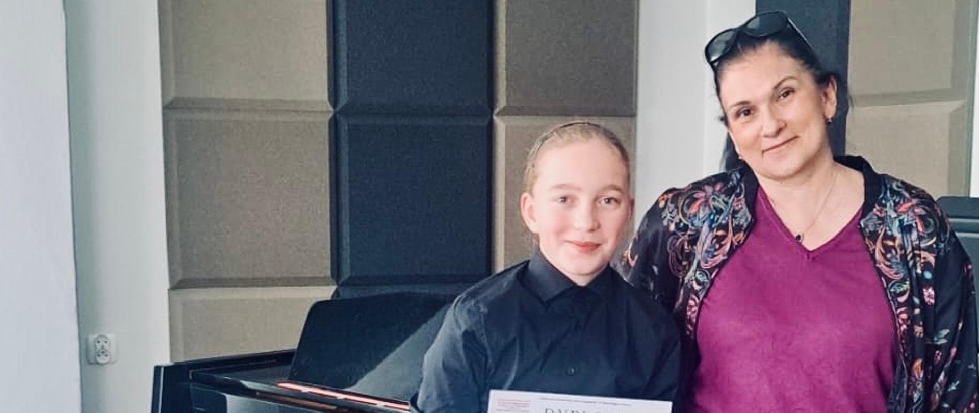 Na zdjęciu widać młodą ucznia Cezarego Sandeckiego stojącą obok nauczycielki Aleksandry Skotarek w sali muzycznej. Za nimi znajduje się fortepian. Uczeń trzyma w rękach dyplom i uśmiecha się do aparatu, a nauczycielka stoi obok z ręką opartą o fortepian. Oboje wyglądają na zadowolonych i dum­nych.