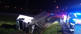 Zdjęcie przedstawia leżący na boku samochód ciężarowy z naczepą. Z prawej strony znajdują się strażacy. Na zewnątrz jest ciemno.
