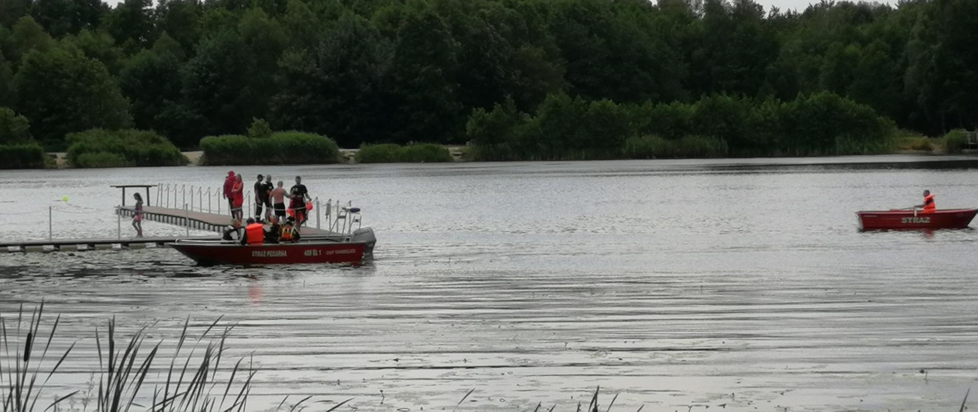 na zdjęciu widoczny zalew na nim dwie łodzie oraz strażacy i ratownicy na pomoście przygotowujący się do ćwiczeń w oddali las