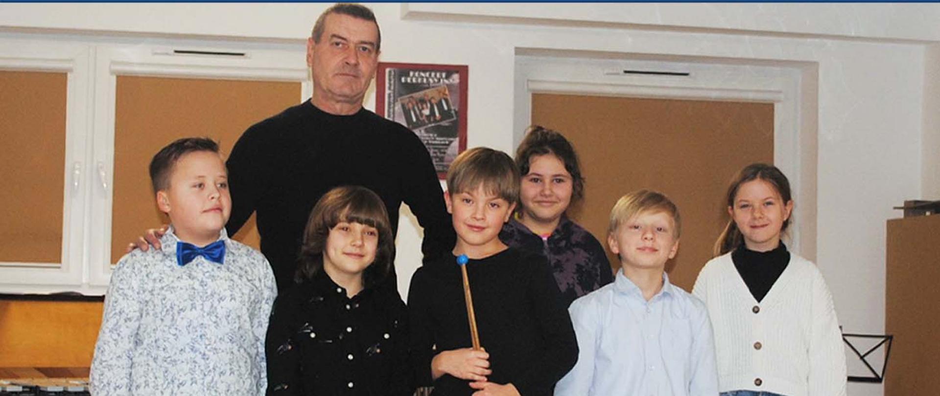 Na zdjęciu nauczyciel klasy perkusji i sześcioro uczniów stoją w pomieszczeniu, w tle okna pomieszczenia.
