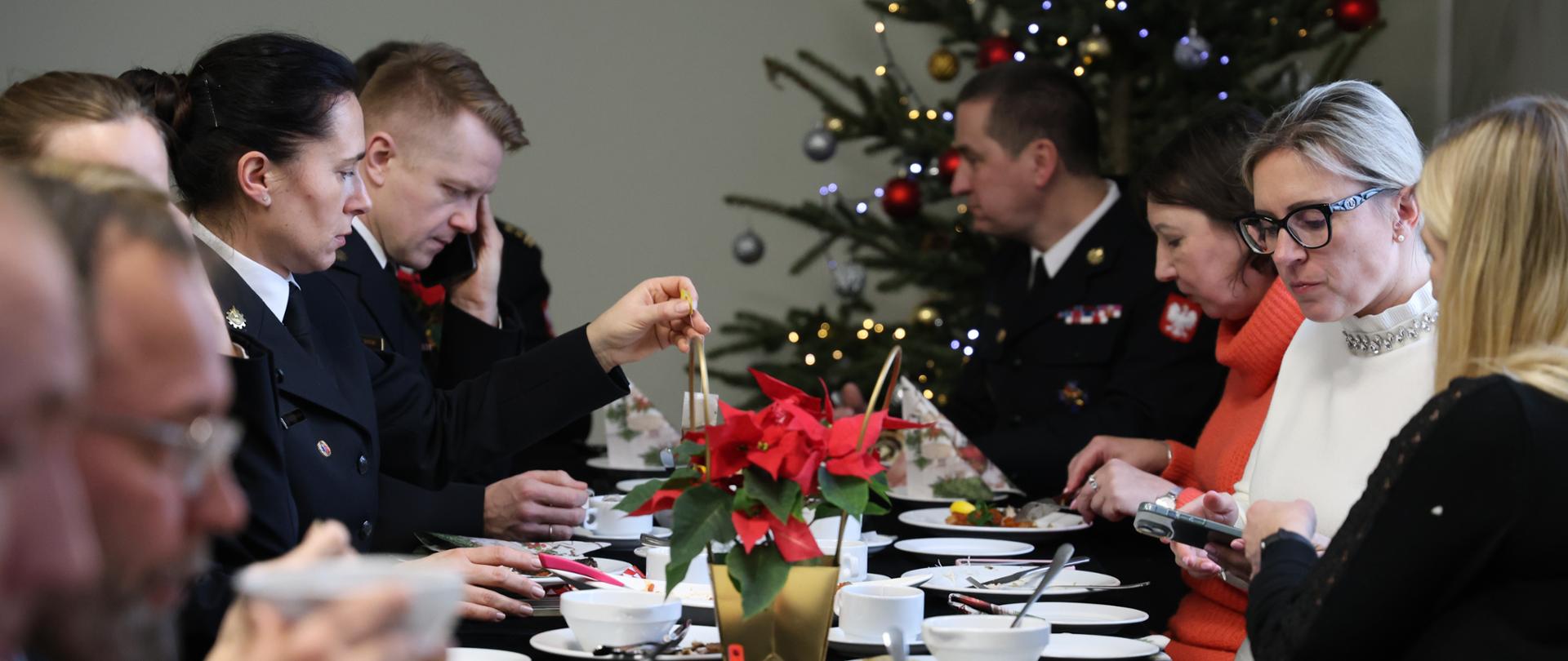 Zdjęcie przedstawia grupę dorosłych osób siedzących przy długim stole podczas uroczystego spotkania. Większość osób ma na sobie ciemne, eleganckie stroje lub mundury. Na stole stoją białe talerze, filiżanki, miseczki oraz półmiski z jedzeniem. Pośrodku stołu znajduje się doniczka z czerwoną gwiazdą betlejemską.
Uczestnicy siedzą blisko siebie, pochylają się nad talerzami, jedzą lub trzymają sztućce. Jedna osoba rozmawia przez telefon, inne skupiają się na posiłku. Atmosfera jest spokojna i poważna.
W tle widoczna jest choinka bożonarodzeniowa ozdobiona światełkami i bombkami, co sugeruje świąteczny charakter spotkania. Pomieszczenie jest jasne, a cała scena wygląda na oficjalne, wspólne świąteczne spotkanie przy stole.