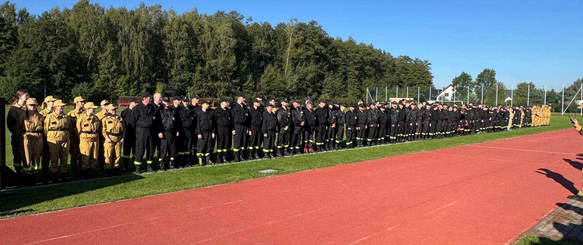 Długi rząd strażaków w mundurach czarnych i beżowych ustawionych wzdłuż czerwonej bieżni, na tle linii drzew i nieba.