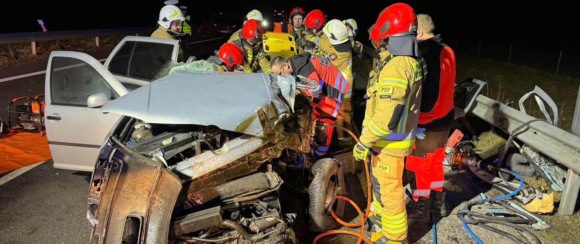 Rozbity pojazd stoi nocą na drodze ekspresowej, dookoła stoją strażacy w czerwonych hełmach i ratownicy medyczni.