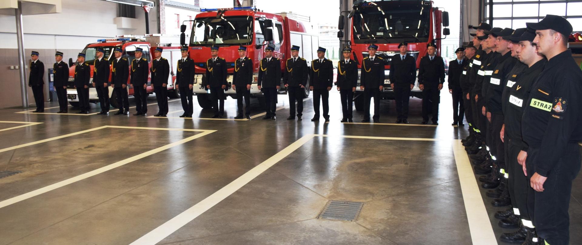 Zbiórka z okazji przejścia na zaopatrzenie emerytalne strażaka. W tle pododdział strażaków w mundurach wyjściowych koloru ciemnogranatowego stojący przed wozami strażackimi koloru czerwonego.