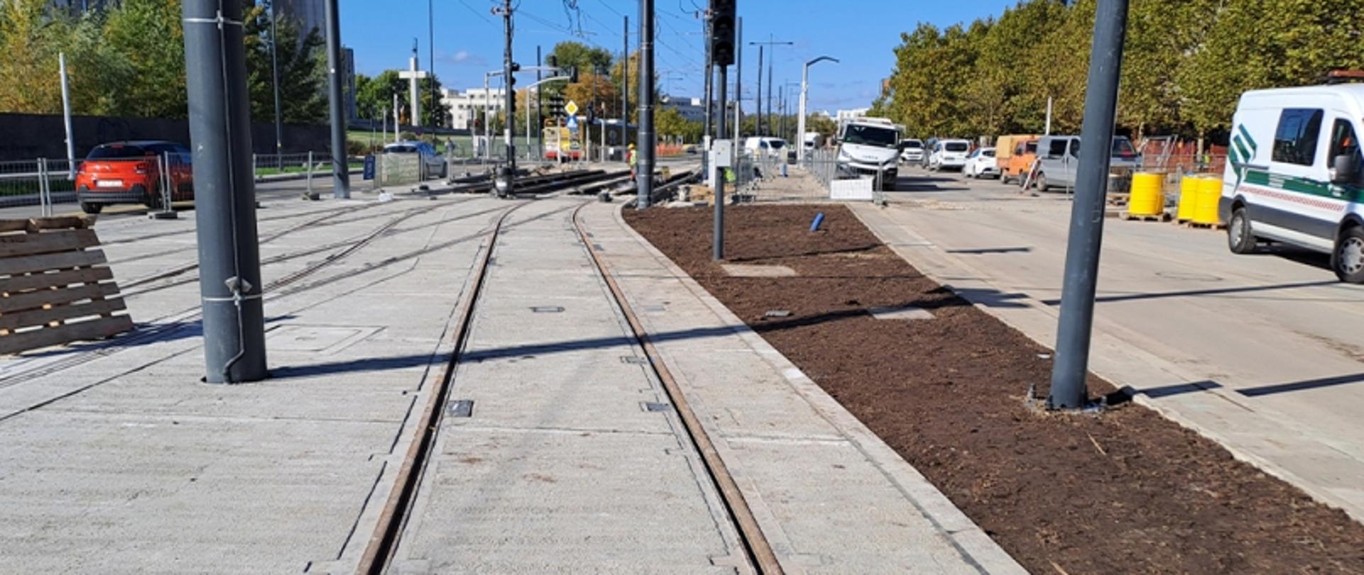 Budowa torowiska tramwajowego z ułożonymi szynami, sprzętem budowlanym i przygotowanym terenem pod nasadzenia; w tle pojazdy i zabudowa miejska.