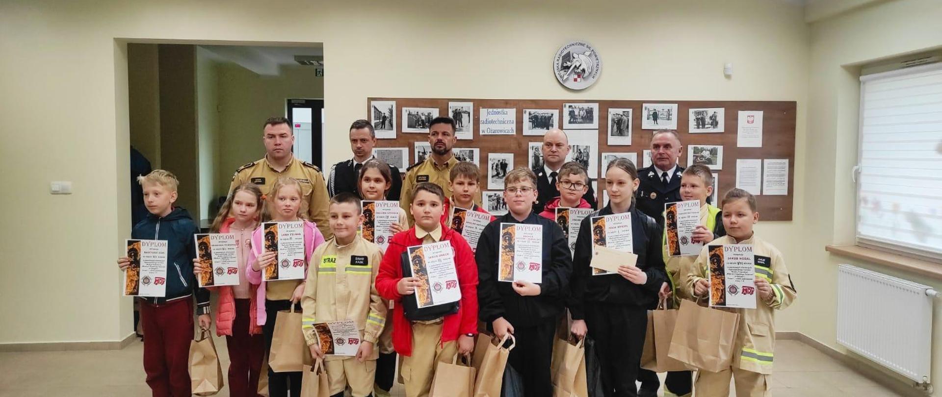 W pierwszym rzędzie stoją dzieci (dziewczynki i chłopcy). W rękach trzymają dyplomy oraz papierowe torby. Za nimi stoją panowie strażacy.