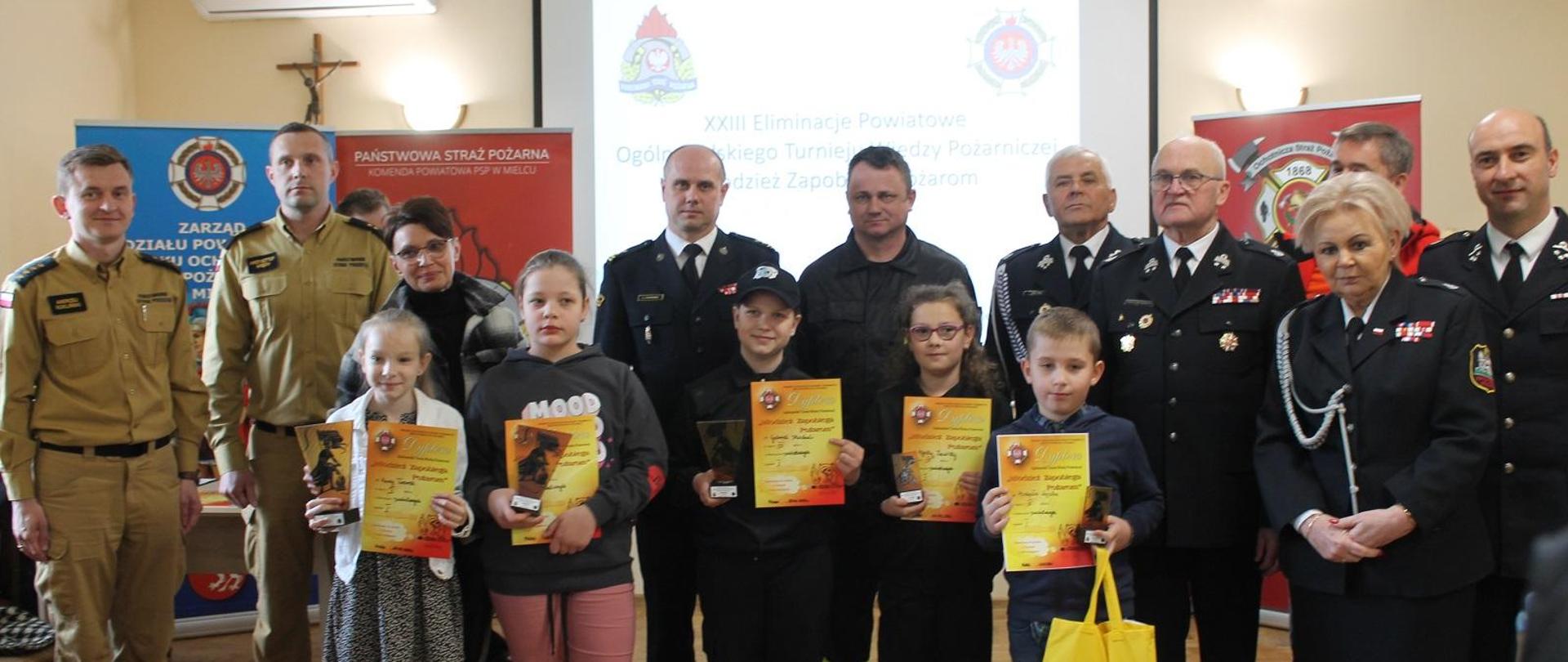 Fotografia wykonana podczas eliminacji powiatowych OTWP, które odbyły się 20 kwietnia 2024 roku w strażnicy jednostki przy ul. Kilińskiego Ochotniczej Straży Pożarnej w Mielcu. Na fotografii w pierwszym rzędzie stoją laureaci konkursu z najmłodszej grupy wiekowej trzymając w rękach nagrody, statuetki i dyplomy. W drugim rzędzie stoją zaproszeni goście oraz komisja oceniająca konkurs z komendy powiatowej Państwowej Straży Pożarnej w Mielcu. Zdjęcie wykonane na tle telebimu i banerów w sali jednostki OSP.