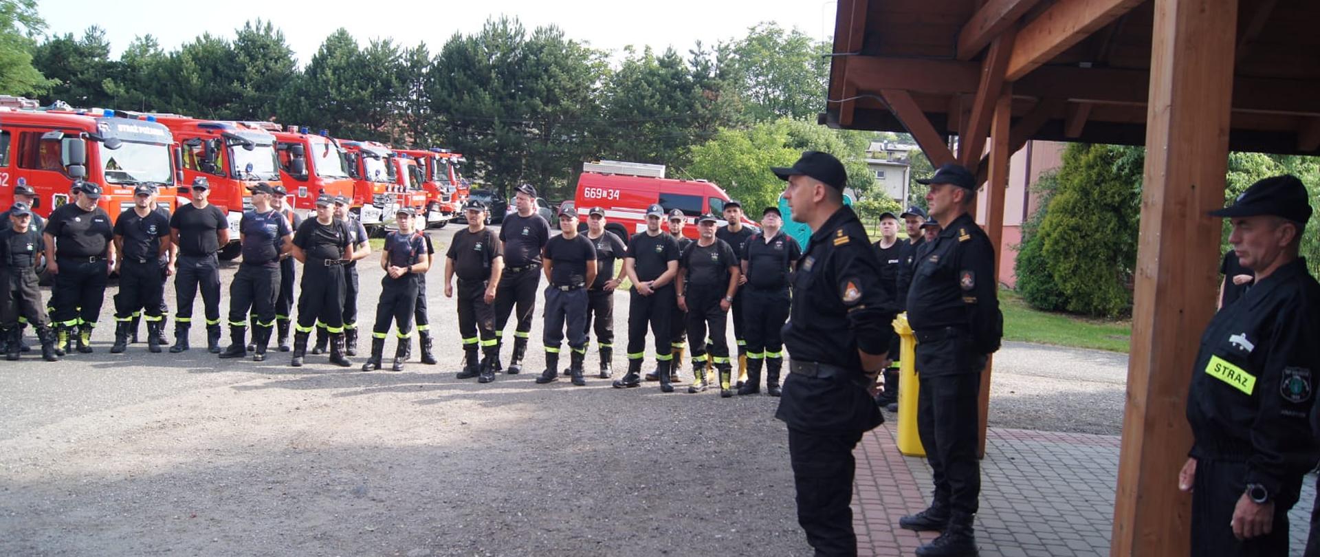 Na zdjęciu strażacy na tle pojazdów pożarniczych podczas zbiórki przed rozpoczęciem ćwiczeń