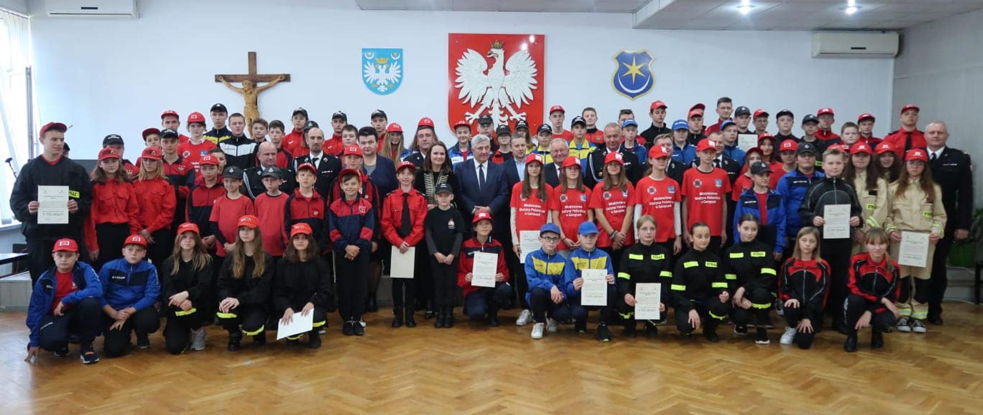 Grupowa fotografia, gdzie w kilku rzędach stoją dzieci, członkowie młodzieżowych drużyn pożarniczych ubrane w różnokolorowe mundurki. W rękach trzymają kartki z tekstem promes. Pośrodku stoi Pani Minister, starosta oraz przedstawiciele samorządu z terenów gmin powiatu przeworskiego. Na zdjęciu w mundurach galowych wodocznych jest dwóch funkcjonariuszy Państwowej Straży Pożarnej