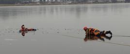 Zdjęcie przedstawia strażaka na desce lodowej podczas ćwiczeń. Jeden ze strażaków znajduje się w wodzie. Kolejny strażak przy pomocy deski lodowej ratuje poszkodowanego. 