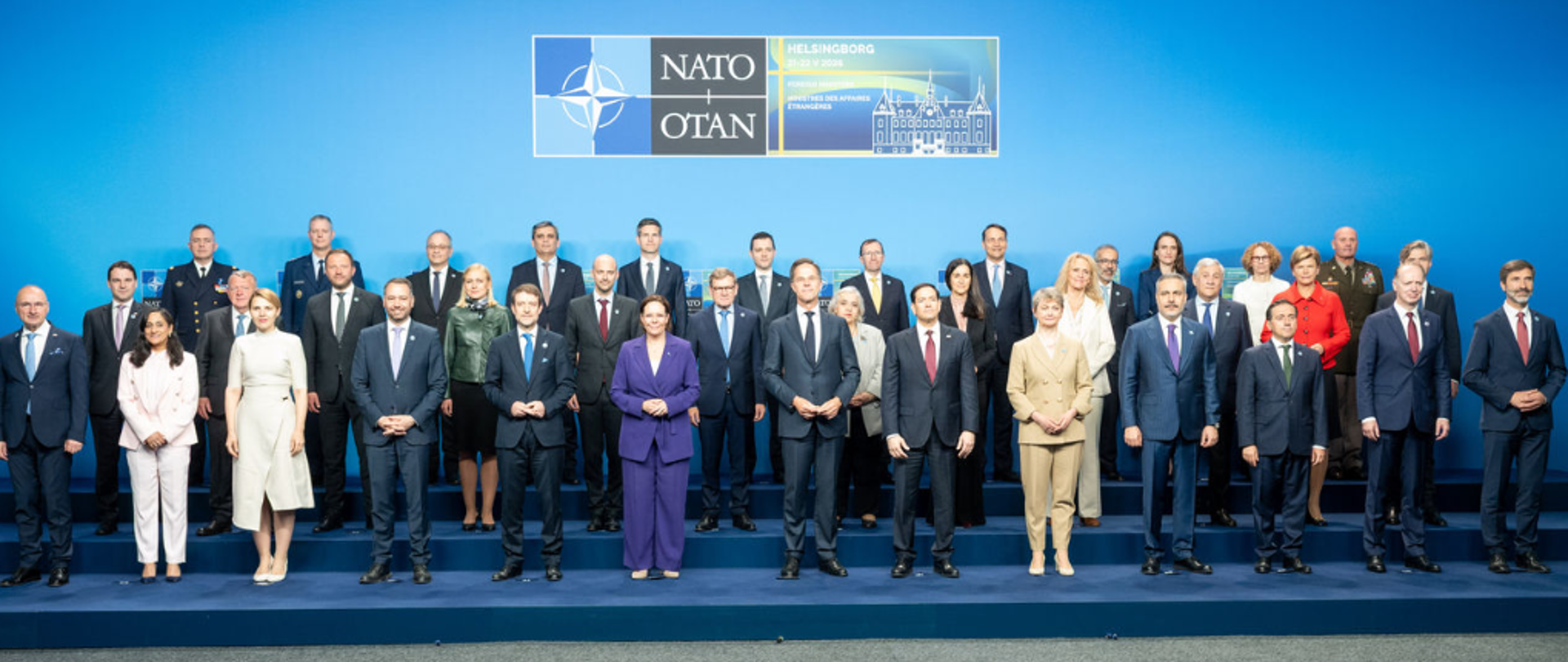 Grupowe zdjęcie uczestników szczytu NATO pozujących do wspólnej fotografii na tle niebieskiej ściany z logo NATO i napisem „Helsingborg”. Politycy i przedstawiciele państw stoją w kilku rzędach, ubrani formalnie – w garnitury i eleganckie stroje.