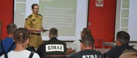 Inauguracja szkolenia podstawowego. Zdjęcie przedstawia świetlicę komendy. Za stołami siedzą uczestnicy szkolenia. Na przeciwko nich, na tle ekranu stoi strażak w mundurze służbowym koloru piaskowego, prowadzący zajęcia. Na ekranach wyświetlana jest prezentacja.