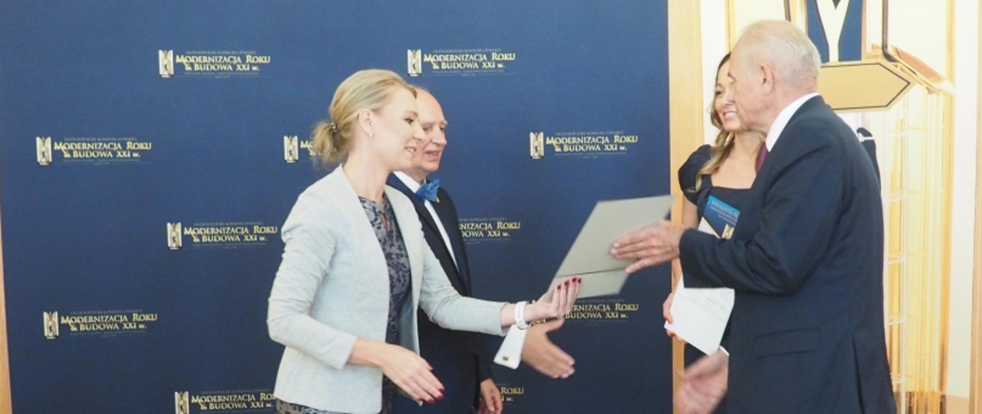 Wręczenie nagrody podczas wydarzenia „Modernizacja Roku & Budowa XXI w.” na tle niebieskiego banera.