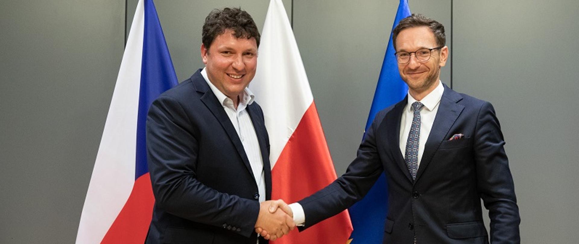 Ministrowie Marian Piecha i Waldemar Buda podają sobie dłonie na tle flag Polski i Czech