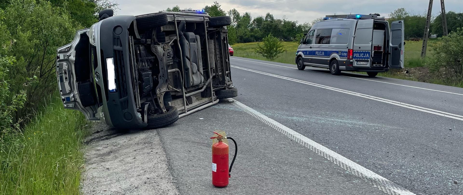Na pierwszym planie gaśnica, na drugim planie samochód leżący na prawym boku. W tle po prawej stronie znajduje się radiowóz policyjny. 