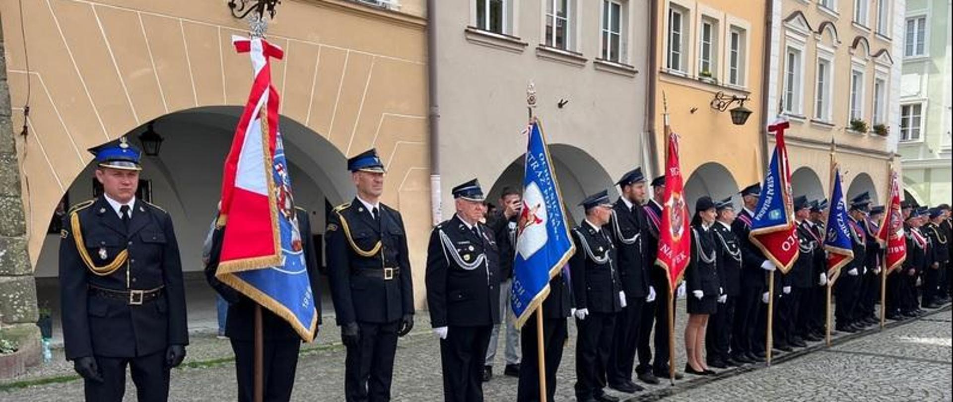 Strażacy stoją na placu ratuszowym i trzymają sztandary
