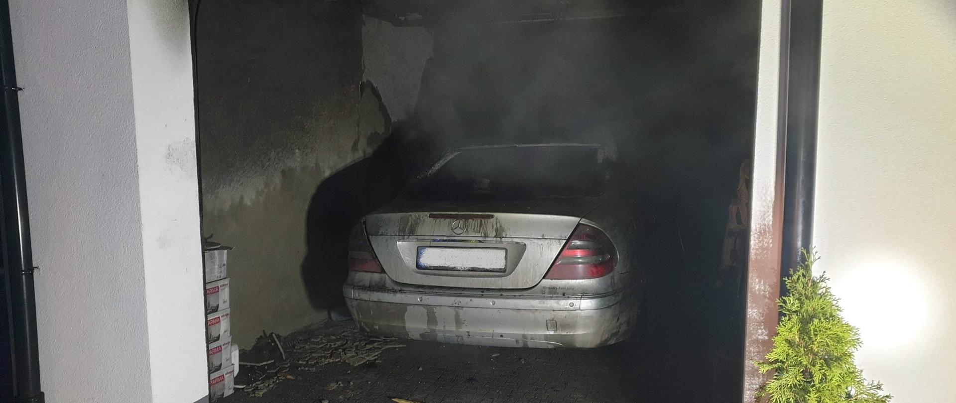 Zdjęcie przedstawia spalony samochód koloru srebrnego, który znajduje się w garażu