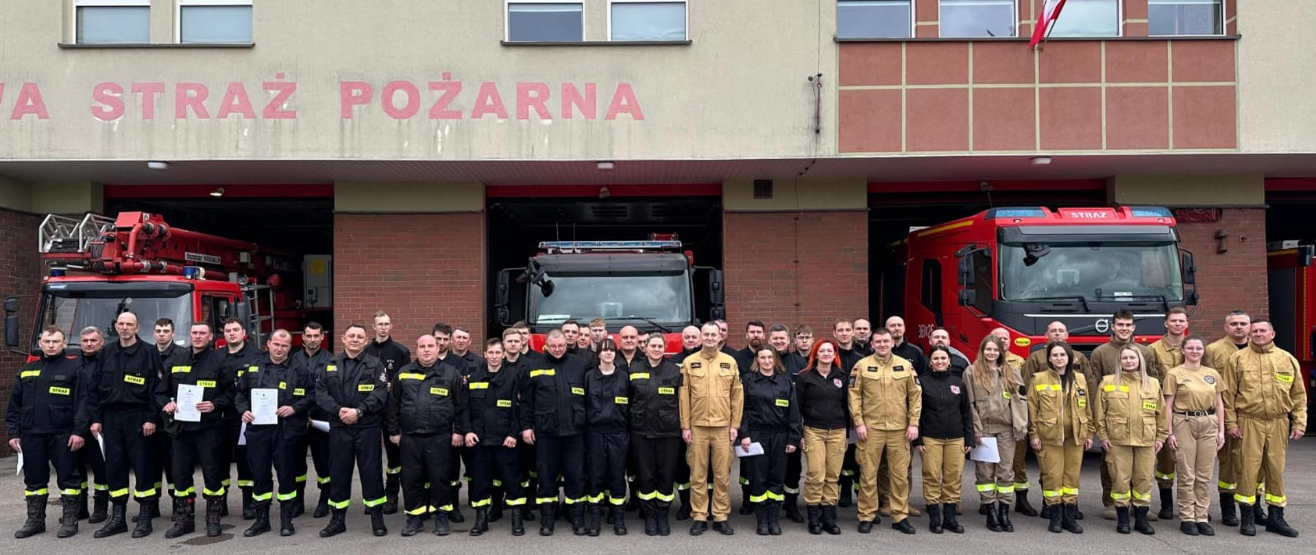 Wspólne zdjęcie absolwentów szkolenia podstawowego strażaka OSP i kadry szkoleniowej. W tle pojazdy ratownicze oraz budynek KM PSP w Łomża.