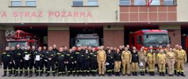Wspólne zdjęcie absolwentów szkolenia podstawowego strażaka OSP i kadry szkoleniowej. W tle pojazdy ratownicze oraz budynek KM PSP w Łomża.