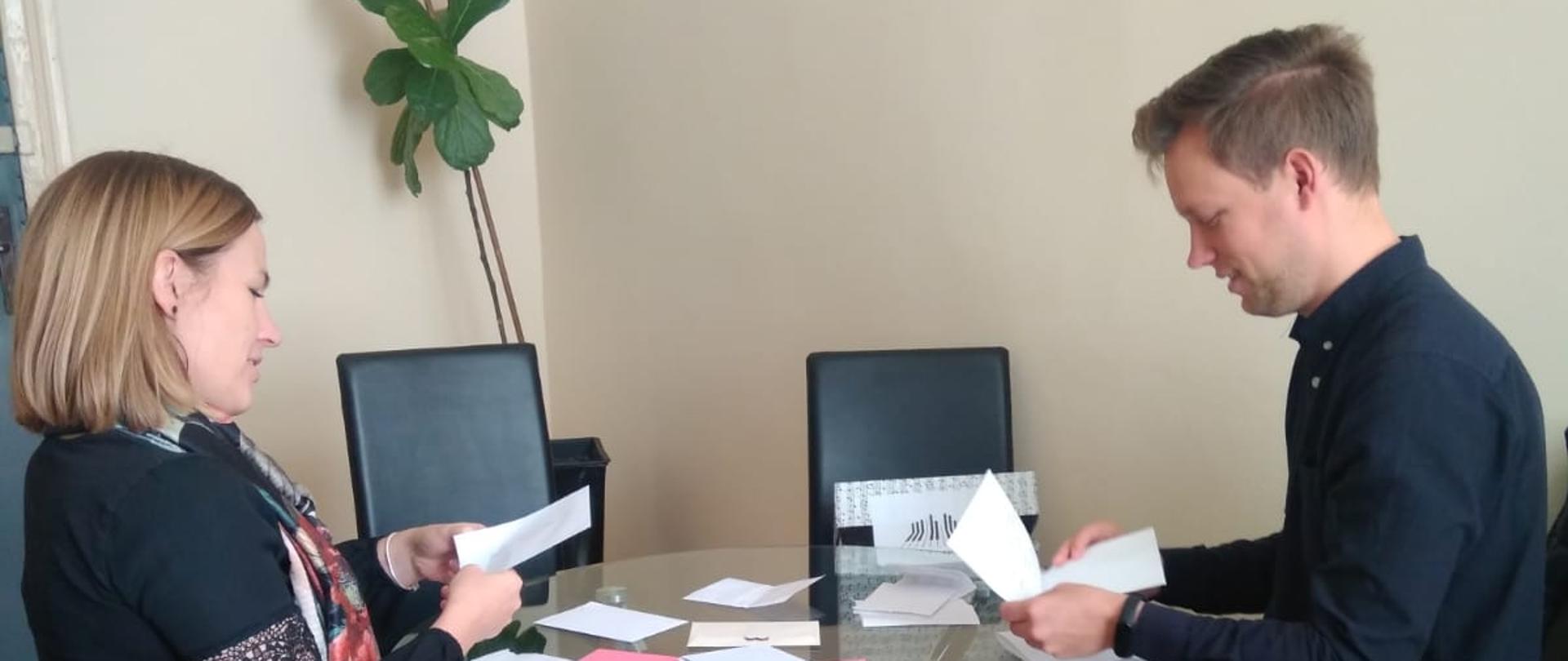 Na zdjęciu w środkowej części widzimy szklany stół, na którym znajduje się wiele kopert i kartek. Stół otacza pięć czarnych krzeseł. Na dwóch z nich, naprzeciwko siebie siedzą dwie postacie odczytujące zapisane na kartkach informacje. W tle znajduje się zielony kwiat.