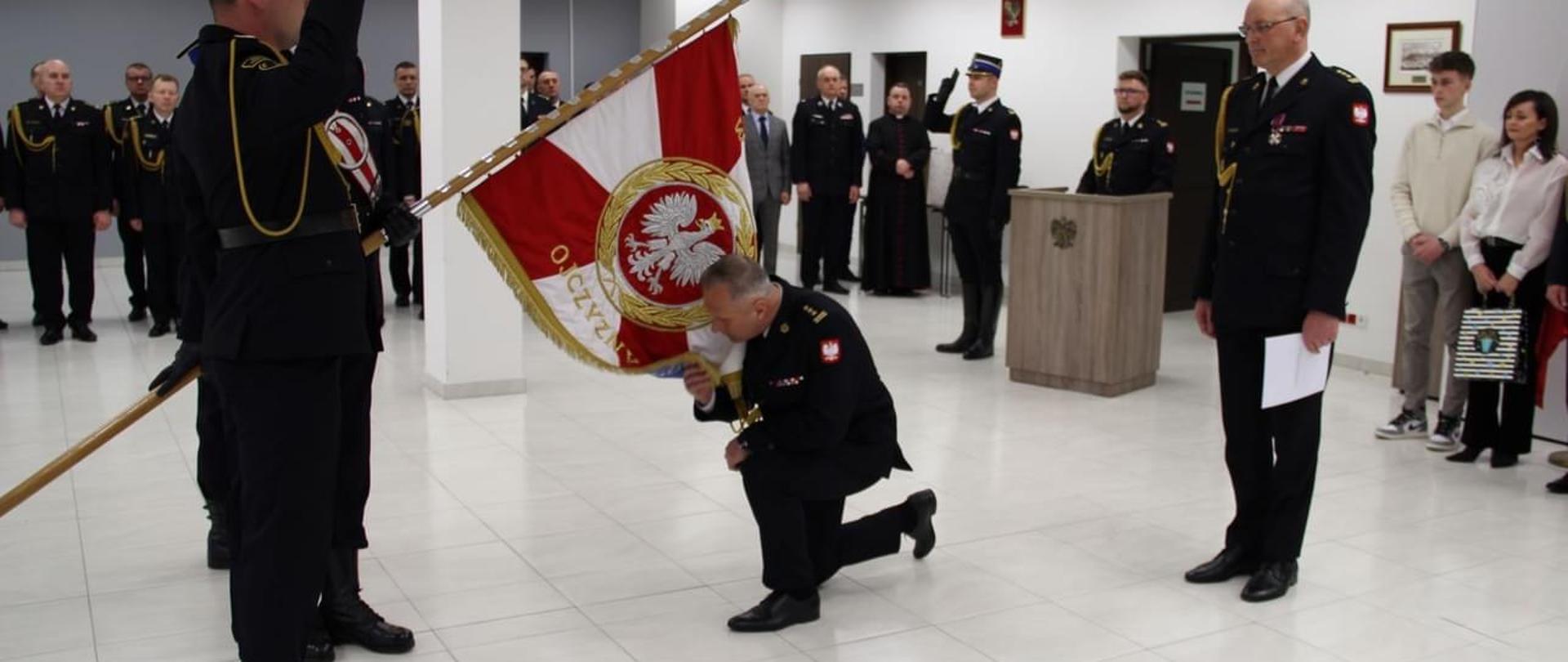 Komendant w obecności przełożonego i załogi, oddaje cześć sztandarowi, klęcząc całuje płat sztandaru. Z lewej strony widok na poczet sztandarowy. W tle goście uroczystości.