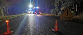 Ustawione na jezdni samochody straży pożarnej obok rozbitego samochodu osobowego. Przed nimi pachołki oraz znak "Wypadek".