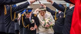 Zdjęcie przedstawia strażaków podczas szpaleru z okazji zaślubin. Strażacy trzymają toporki w rękach. Mają założone również maseczki. Pod toporkami przechodzi para młoda. Pan młody ubrany w mundur galowy PSP zaś panna młoda w białą suknię, welon i bukiet kwiatów.