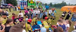 Zdjęcie przedstawia pokaz pierwszej pomocy prowadzony przez strażaków na otwartej przestrzeni. W centrum kadru kilku strażaków w mundurach i kaskach udziela pomocy leżącemu na ziemi człowiekowi-manekinowi. Wokół nich zgromadziła się duża grupa gapiów, w tym wiele dzieci, obserwujących akcję. W tle widoczne są duże, kolorowe nadmuchiwane zamki i zjeżdżalnie, a także pomarańczowy namiot. Scena rozgrywa się na trawiastym terenie. W prawym dolnym rogu zdjęcia widnieje napis "25 czerwca 2025 10:56 Komenda Powiatowa PSP w Białobrzegach".
