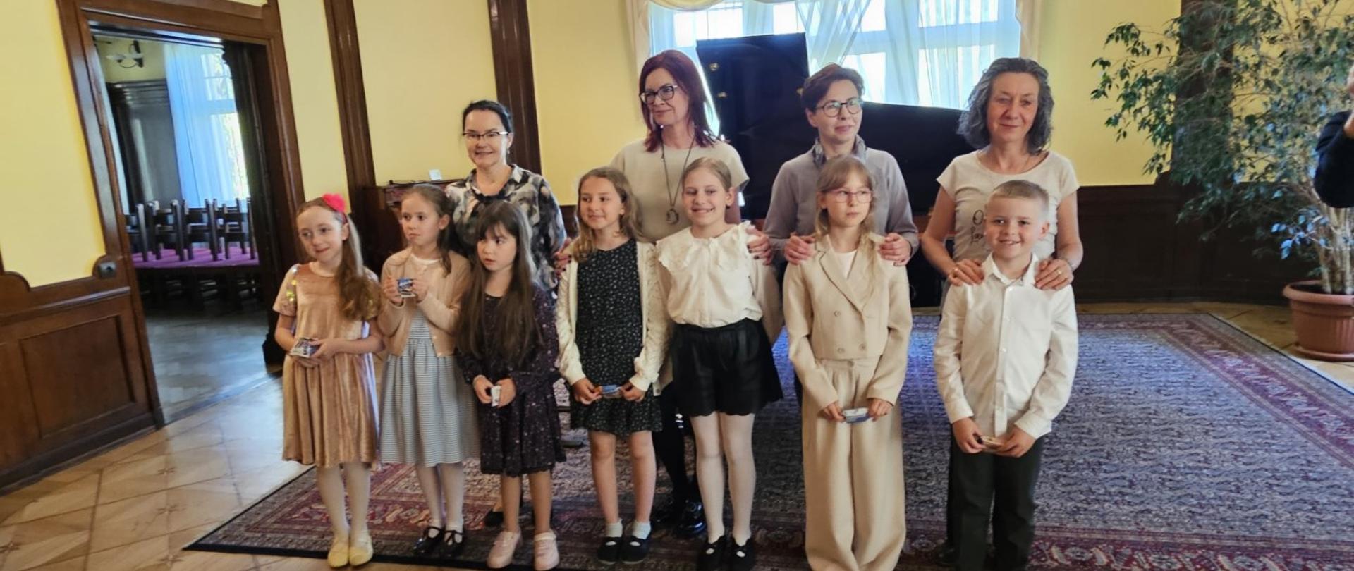 Zdjęcie kolorowe. Zdjęcie przedstawia grupę dzieci (sześć dziewczynek i jednego chłopca) oraz ich nauczycielki (cztery panie stojące za dziećmi), wszyscy są odświętnie ubrani. W tle znajduje się skrzydło fortepianu, duże okna oraz stylowe ściany w żółtym kolorze. Atmosfera zdjęcia emanuje dumą po udanym koncercie. 