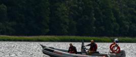 Łódź ratunkowa Straży Pożarnej z trzema strażakami w kamizelkach ratunkowych płynie po jeziorze. Na łodzi widoczny jest napis „STRAŻ” i silnik marki Honda. W tle znajduje się pas zieleni i las.