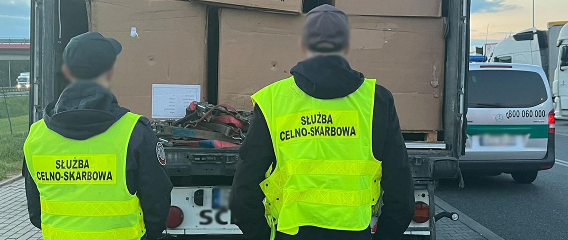 Dwie osoby w kamizelkach z napisem „Służba Celno‑Skarbowa” stoją przy otwartej przestrzeni ładunkowej ciężarówki wypełnionej kartonami.