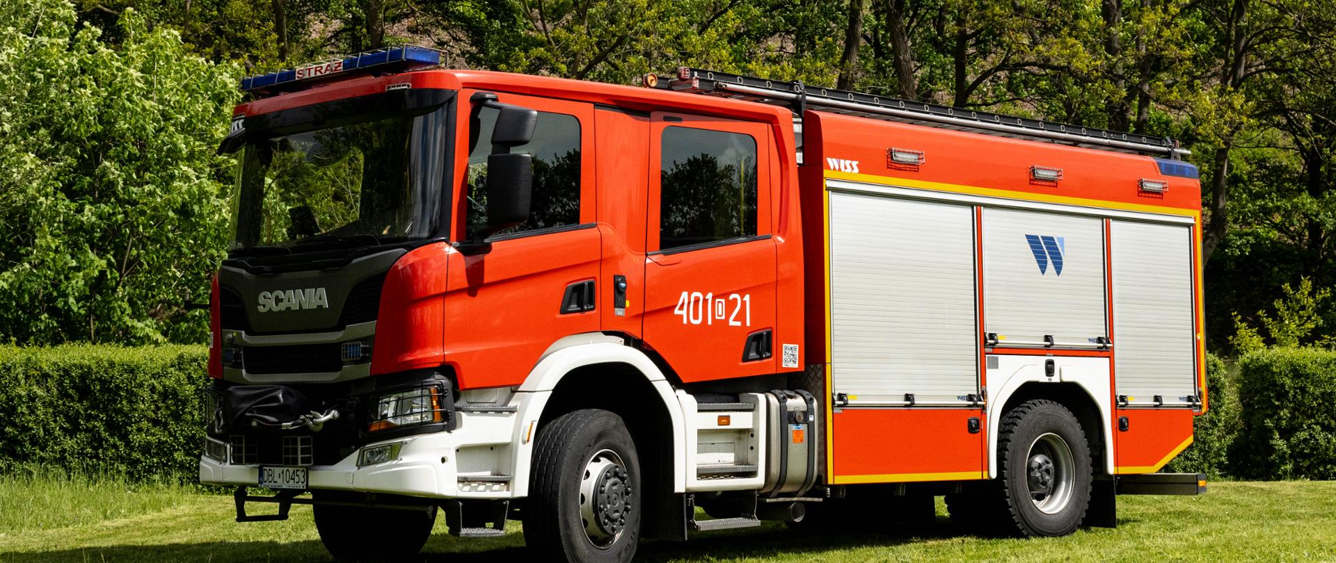 Średni samochód ratowniczo-gaśniczy marki Scania P360 w kolorze czerwonym z białymi nadkolami i zderzakiem. Na drzwiach kabiny znajduje się numer operacyjny „401[D]21”, a na bocznych, srebrnych żaluzjach widoczne jest niebieskie logo „WISS”. Wóz zaparkowany jest na trawie przed ścianą lasu.