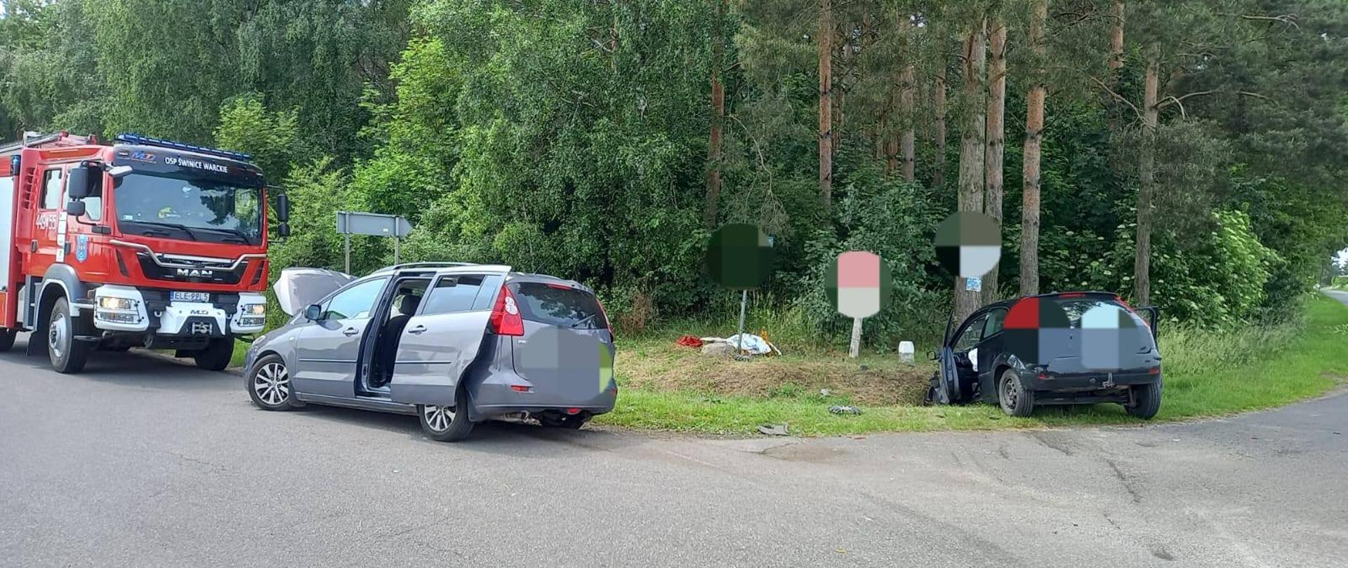 Na zdjęciu widać wypadek drogowy, w którym uczestniczyły dwa samochody osobowe. Jeden z samochodów, jest koloru srebrnego i znajduje się na jezdni. Drzwi boczne pojazdu są otwarte, a maska uniesiona. Drugi czarny samochód wypadł z drogi i znajduje się na poboczu – trawie. Samochód ma widoczne uszkodzenia oraz otwarte przednie drzwi od strony kierowcy. Na miejscu widoczny jest wóz straży pożarnej. W tle znajdują się drzewa oraz znaki drogowe. Na poboczu znajdują się porozrzucane elementy pojazdów.