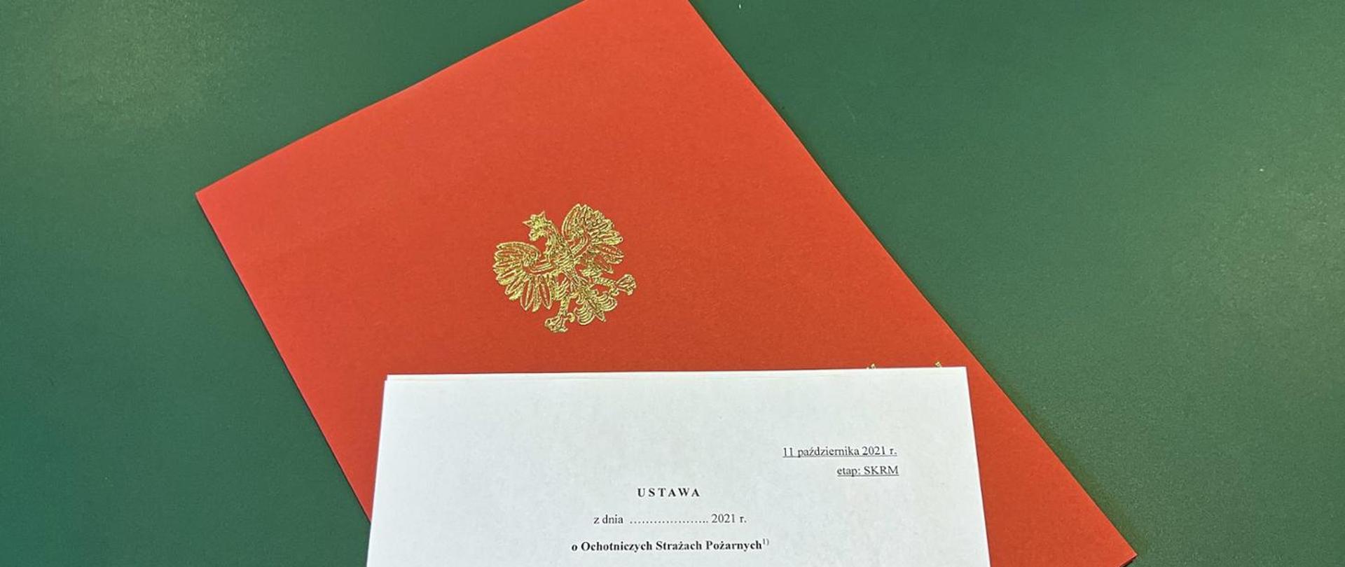 Biała kartka z tytułem dokumentu ułożona na czerwonej teczce papierowej z orłem, pod spodem zielone tło