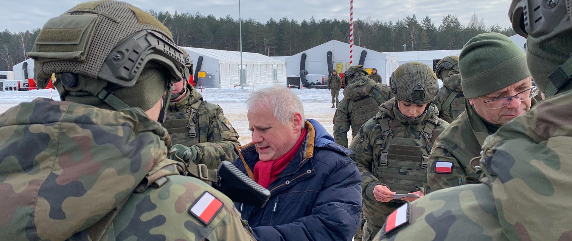 Mężczyzna w granatowej kurtce rozmawia z żołnierzami, ma poważną minę 