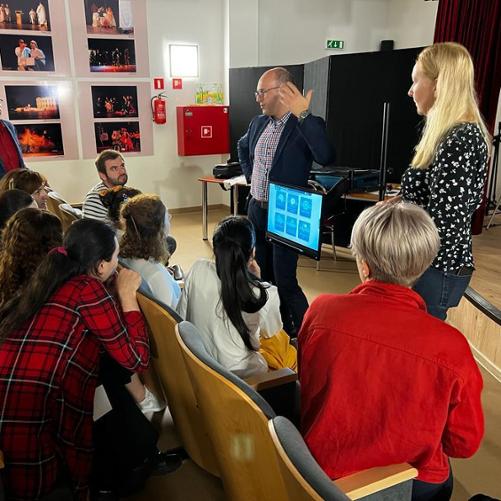 Kobieta i mężczyzna stoją w sali przy monitorze, mężczyzna coś wyjaśnia grupie osób zgromadzonych przed nimi.