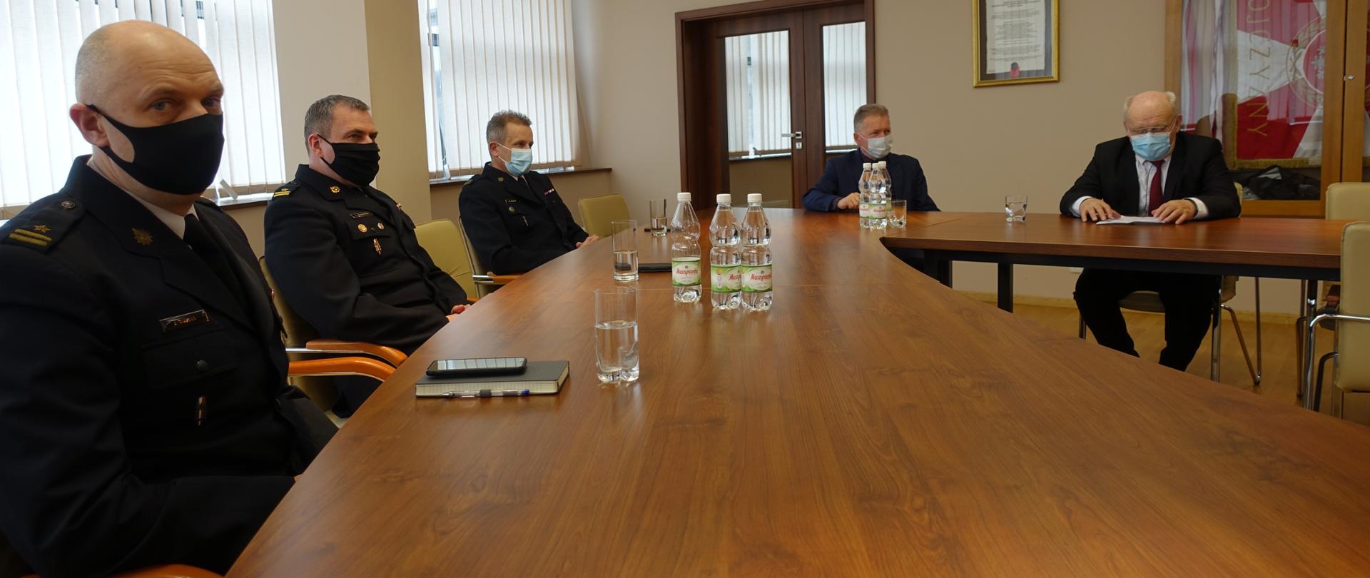 Kadra kierownicza KM PSP Nowy Sącz oraz goście w maseczkach z zachowaniem odległości siedzą przy stole w sali narad.