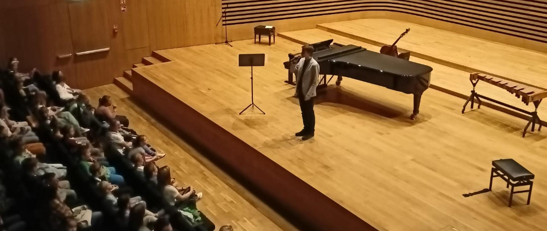 Mężczyzna stoi na estradzie sali koncertowej PSM w Jastrzębiu-Zdroju i mówi do mikrofonu, za Nim stoi fortepian i ksylofon, po lewej stronie widać zapełnioną widownię.