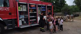 Dzieci zgromadzone przed otwartymi skrytkami samochodu gaśniczego, przy nich strażak prowadzący prelekcje. 