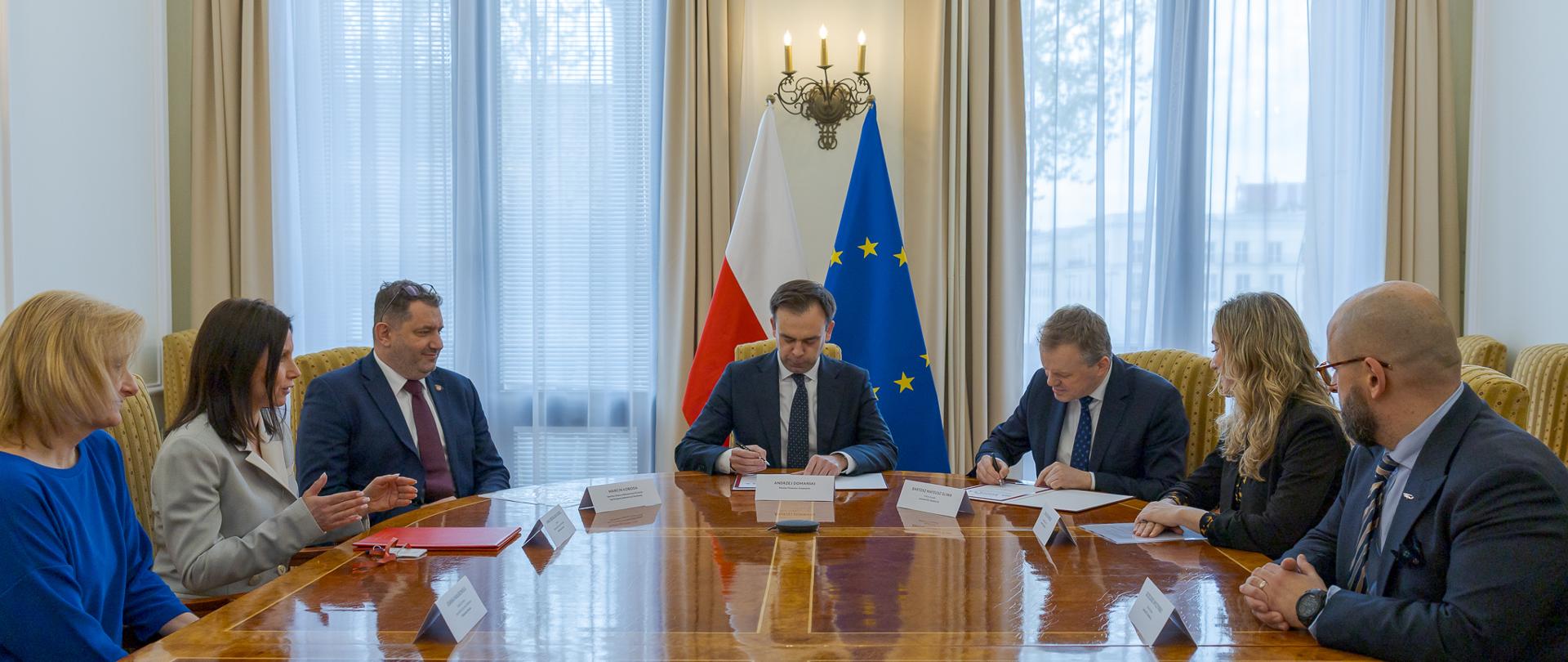 Uczestnicy spotkania w tym Minister Finansów i Gospodarki Andrzej Domański siedzą przy stole konferencyjnym w sali z flagą Polski i Unii Europejskiej.