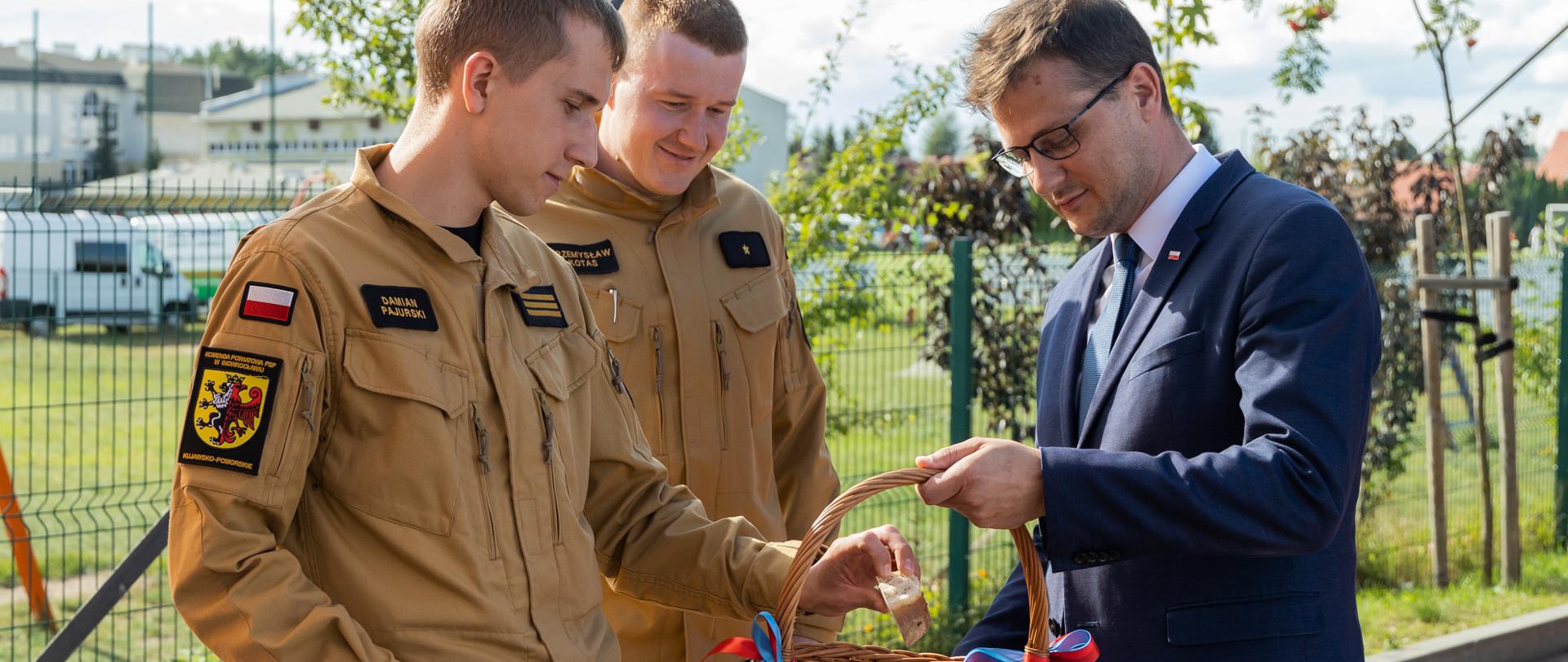 Oficjalne spotkanie plenerowe. Mężczyzna w ciemnym garniturze i okularach trzyma duży wiklinowy kosz przewiązany kolorowymi wstążkami i rozmawia z dwoma młodymi strażakami w jasnobrązowych mundurach. Jeden ze strażaków sięga dłonią do wnętrza kosza.